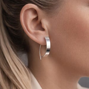 Puede incluir: Primer plano de un pendiente de aro plateado. El pendiente tiene una parte delantera plana y rectangular y un alambre fino y curvado que atraviesa la oreja. El pendiente es brillante y reflectante, y el diseño general es minimalista y moderno.