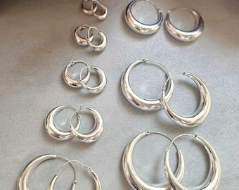 May include: A collection of ten silver-toned hoop earrings arranged in five pairs of varying sizes. Each pair features a smooth, polished, rounded metal design with a thin wire post closure, displayed in a graduated layout from smallest to largest.