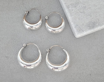 May include: Six silver hoop earrings are displayed. The earrings are arranged in three pairs, each with a rounded, crescent shape. The silver hoops have a polished, reflective surface. The earrings are presented on a grey surface.