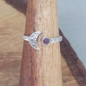 May include: A silver ring with a crescent moon design and a textured finish, set with a small, round, purple gemstone. The ring is presented on a wooden ring holder, highlighting its celestial theme and intricate details.