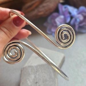 May include: A silver arm cuff bracelet with spiral ends. The open cuff design features two decorative spirals, one at each end. The bracelet is shown against a blurred background with hints of purple and brown.