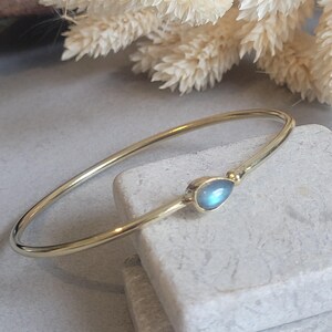 May include: A thin, polished gold-tone metal bangle bracelet featuring a single, teardrop-shaped cabochon gemstone. The stone displays a soft, iridescent blue glow, set securely within a smooth metal bezel at the centre of the circular band.