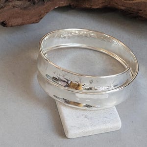 May include: Two silver bangles with a hammered finish. The bracelets are stacked, with one overlapping the other. The jewellery is displayed on a small, white, rectangular stone, with a blurred wooden element in the background.