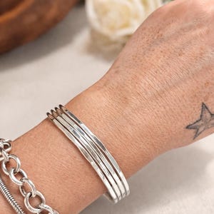 Puede incluir: Un brazalete plateado con cuatro bandas paralelas. También se ve un segundo brazalete plateado con un diseño de cadena. Los brazaletes se llevan en una muñeca con un tatuaje de estrella.