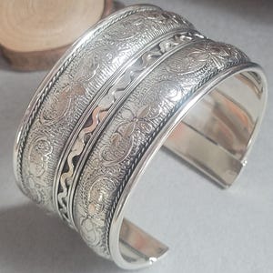 May include: A wide silver cuff bracelet with intricate floral and scrollwork patterns. The bracelet features a detailed design with a textured surface and a polished finish. The open-ended design allows for easy wear.