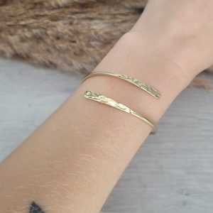 May include: A gold hammered metal bangle bracelet with a simple, open design. The bracelet is made of two thin pieces of metal that are joined together at the ends.