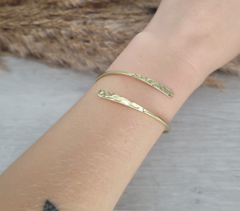 May include: A gold hammered metal bangle bracelet with a simple, open design. The bracelet is made of two thin pieces of metal that are joined together at the ends.