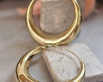 May include: A pair of polished gold-tone hoop earrings featuring a thick, rounded, crescent-shaped design. The earrings have a smooth, reflective surface and a thin wire post closure, showcasing a bold, curved silhouette in a bright metallic finish.