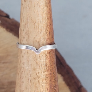 May include: A silver ring with a V-shaped design. The ring is displayed on a wooden ring holder. The ring's simple design makes it suitable for everyday wear. The ring is a piece of jewellery.