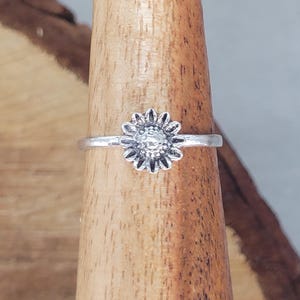 Könnte beinhalten: Ein silberfarbener Ring mit einem detaillierten Sonnenblumen-Design. Der Ring hat ein schmales Band, und die Sonnenblume weist aufwendige Blütenblattdetails um einen strukturierten Kern auf. Der Ring wird auf einer Holzoberfläche präsentiert.