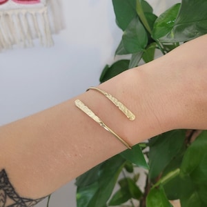 May include: A gold hammered metal bracelet with a simple, open design. The bracelet is made of two thin, flat pieces of metal that are joined together at the ends. The metal has a hammered texture, which gives it a unique and rustic look.