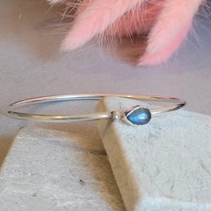 May include: A thin, polished silver bangle bracelet featuring a single teardrop-shaped labradorite gemstone with a blue iridescent flash. The bracelet includes a small, smooth silver bead accent positioned next to the gemstone setting.