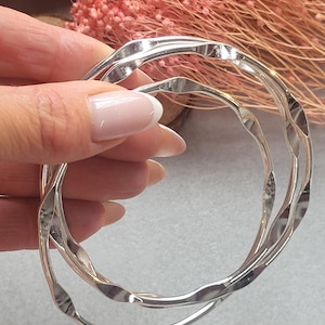 May include: Two circular silver bangles featuring a continuous wavy, rippled texture along the entire circumference. The metal surface is polished and reflective, creating a series of light and dark highlights along the undulating edges of the rings.