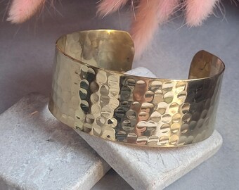 May include: A wide, polished gold-tone metal cuff bracelet featuring a hammered, dimpled texture across its entire outer surface. The bracelet has an open-ended design, allowing for adjustment, and displays a reflective, metallic finish throughout.