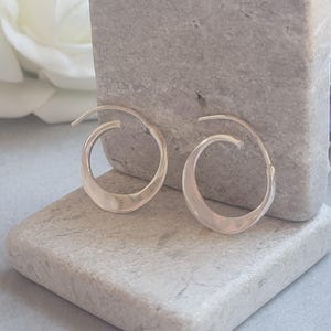 Puede incluir: Un par de pendientes de aro dorados con un diseño abierto único. Los pendientes se exhiben sobre una superficie de piedra gris claro y texturizada, con una flor blanca en el fondo. Los pendientes tienen forma de media luna curvada.