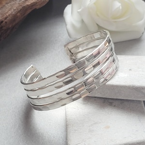 May include: A silver cuff bracelet with a hammered texture. The bracelet has four bands and a slight curve. The metal has a polished finish, reflecting light. The bracelet is displayed on a light grey surface.