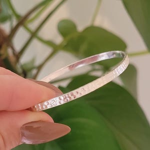 Può includere: Un braccialetto a cerchio martellato in argento. Il braccialetto presenta una superficie strutturata con piccole rientranze uniformemente distanziate. Il braccialetto è tenuto contro uno sfondo di fogliame verde, con una messa a fuoco morbida sullo sfondo.