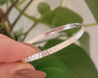May include: A silver hammered bangle bracelet. The bracelet has a textured surface with small, evenly spaced indentations. The bangle is held against a backdrop of green foliage, with a soft focus in the background.