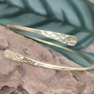 May include: A gold-colored, hammered-finish cuff bracelet. The open-ended design features textured, flattened ends. The bracelet is displayed on a piece of driftwood, with a green leaf in the background. Jewelry.