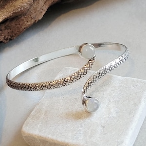 May include: A silver-toned cuff bracelet with a textured, patterned design. The bracelet features two round, light-colored stones at each end. The bracelet is displayed on a light gray stone surface.