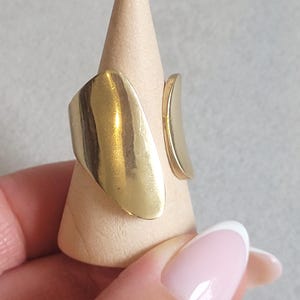 May include: A gold-coloured ring with an abstract design. The ring features a large, curved plate and a smaller, curved piece. It is displayed on a wooden cone-shaped stand.