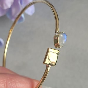 May include: A gold-toned open cuff bracelet featuring a smooth, polished rectangular metal plate on one end and a single, small, iridescent moonstone cabochon set in a gold-toned bezel on the opposite end of the thin, circular metal band.