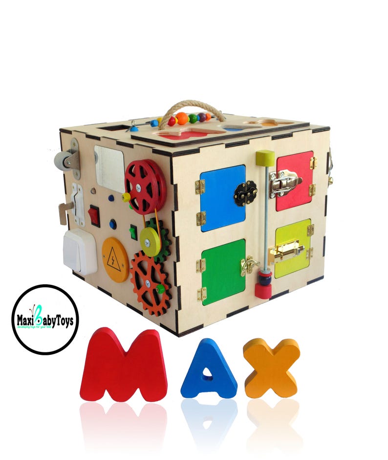 May include: Wooden activity cube with a variety of interactive elements, including gears, latches, a mirror, and a light switch. The cube is painted in bright colors and has a handle on top. The letters MAX are spelled out in colorful wooden blocks in front of the cube.