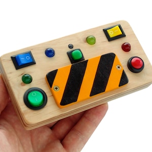 Puede incluir: Un tablero sensorial de madera con varios botones de colores, interruptores y una señal de peligro a rayas negras y amarillas. El tablero presenta botones verdes, azules, amarillos y rojos, e interruptores verdes y azules. El tablero se sostiene en una mano.