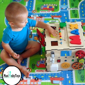 May include: A wooden activity cube with various textures and activities for toddlers. The cube features a sliding door, a spinning gear, a bell, a shape sorter, and a bead maze. The cube is painted in bright colors and has the letters "MAX" on one side. A young child is playing with the cube.