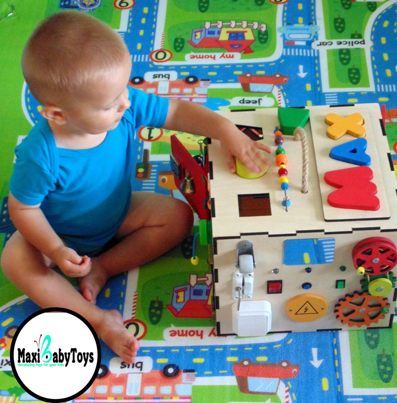 May include: A wooden activity cube with various textures and activities for toddlers. The cube features a sliding door, a spinning gear, a bell, a shape sorter, and a bead maze. The cube is painted in bright colors and has the letters "MAX" on one side. A young child is playing with the cube.