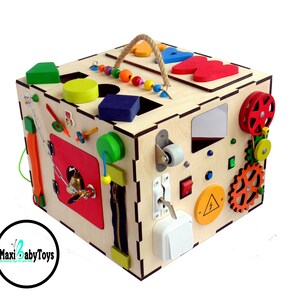 May include: A wooden activity cube with a variety of textures and activities for babies and toddlers. The cube features a zipper, a bell, a latch, a wheel, gears, a mirror, and more. The cube is painted in bright colors and has a rope handle for easy carrying.