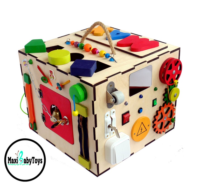 May include: A wooden activity cube with a variety of textures and activities for babies and toddlers. The cube features a zipper, a bell, a latch, a wheel, gears, a mirror, and more. The cube is painted in bright colors and has a rope handle for easy carrying.