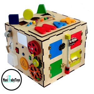May include: Wooden activity cube with various textures and activities for toddlers, including a sliding door, a latch, a gear system, a wheel, and a shape sorter. The cube is painted in bright colors and has the letters "MAX" on the top.