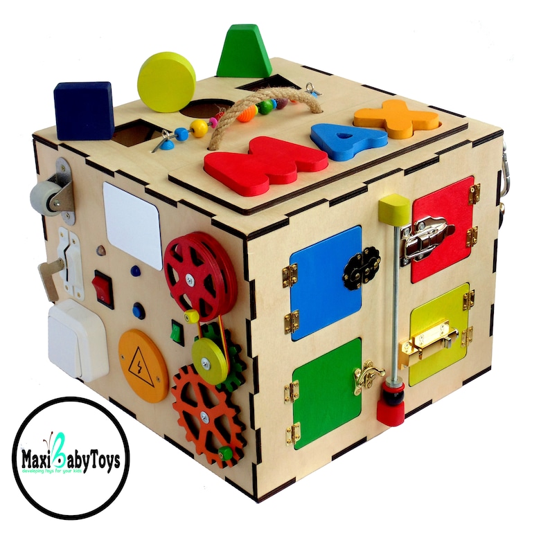 May include: Wooden activity cube with various textures and activities for toddlers, including a sliding door, a latch, a gear system, a wheel, and a shape sorter. The cube is painted in bright colors and has the letters "MAX" on the top.