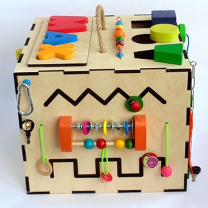 May include: Wooden activity cube with a variety of textures and activities for children. The cube features a bead maze, a shape sorter, a sliding bead track, a latch, a bell, and a maze. The cube is painted in bright colors and has a natural wood finish.