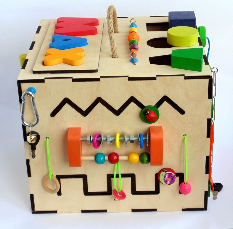 May include: Wooden activity cube with a variety of textures and activities for children. The cube features a bead maze, a shape sorter, a sliding bead track, a latch, a bell, and a maze. The cube is painted in bright colors and has a natural wood finish.