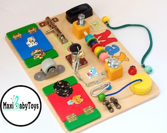 Puede incluir: Un tablero de actividades de madera con varias texturas y actividades para niños pequeños. Cuenta con una puerta azul con bisagras, un pestillo, una rueda, una cadena, una llave, un laberinto de cuentas, un interruptor de palanca y una etiqueta con el nombre "Max".