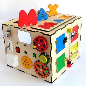 May include: A wooden activity cube with various features for children to explore. It has a sliding door, a gear system, a light switch, a knob, and a small door with a latch. The cube is painted in bright colors, including red, yellow, blue, green, and orange. The top of the cube has the letters "MAX" cut out of it.