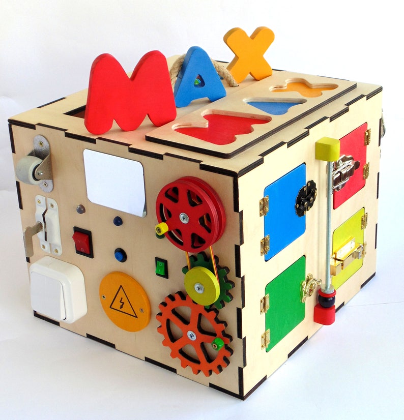 May include: A wooden activity cube with various features for children to explore. It has a sliding door, a gear system, a light switch, a knob, and a small door with a latch. The cube is painted in bright colors, including red, yellow, blue, green, and orange. The top of the cube has the letters "MAX" cut out of it.