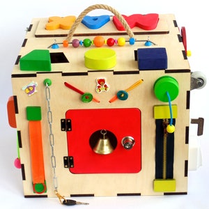 May include: Wooden activity cube with a variety of textures and activities for toddlers. Features include a zipper, a chain, a latch, a bell, a spinning wheel, and a shape sorter. The cube is painted in bright colors, including red, green, blue, yellow, and orange.