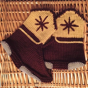 May include: A pair of handmade knitted slippers in shades of brown and tan. The slippers feature a starburst design on the tan upper portion and a suede sole. The knitted material is a dark brown, and the slippers appear cozy and warm.