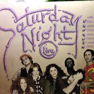 May include: A vintage vinyl record album cover featuring a purple and white graphic of the words "Saturday Night Live" with a group of people standing in front of a white background. The album cover lists the cast members: Dan Aykroyd, John Belushi, Chevy Chase, Jane Curtin, Garrett Morris, Laraine Newman, and Gilda Radner.