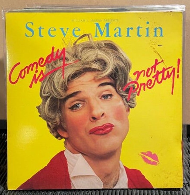 May include: A yellow background with the text "Steve Martin" in black and "Comedy is not pretty!" in red. A man with blonde hair is wearing a red jacket and a white shirt. He is looking at the camera with a serious expression.