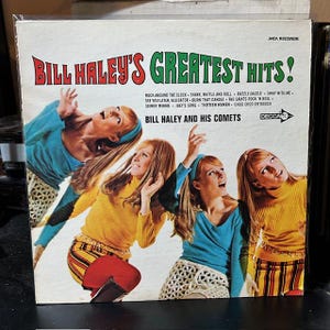 May include: Vintage vinyl record album cover for "Bill Haley's Greatest Hits!" featuring a photo of four women in colorful outfits. The album title is in red and green, with song titles listed below. The artist is "Bill Haley and His Comets."