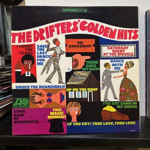 May include: Vintage vinyl record album cover for "The Drifters' Golden Hits." The cover features colorful illustrations and text, including song titles like "On Broadway" and "Under the Boardwalk." The album art has a retro aesthetic.