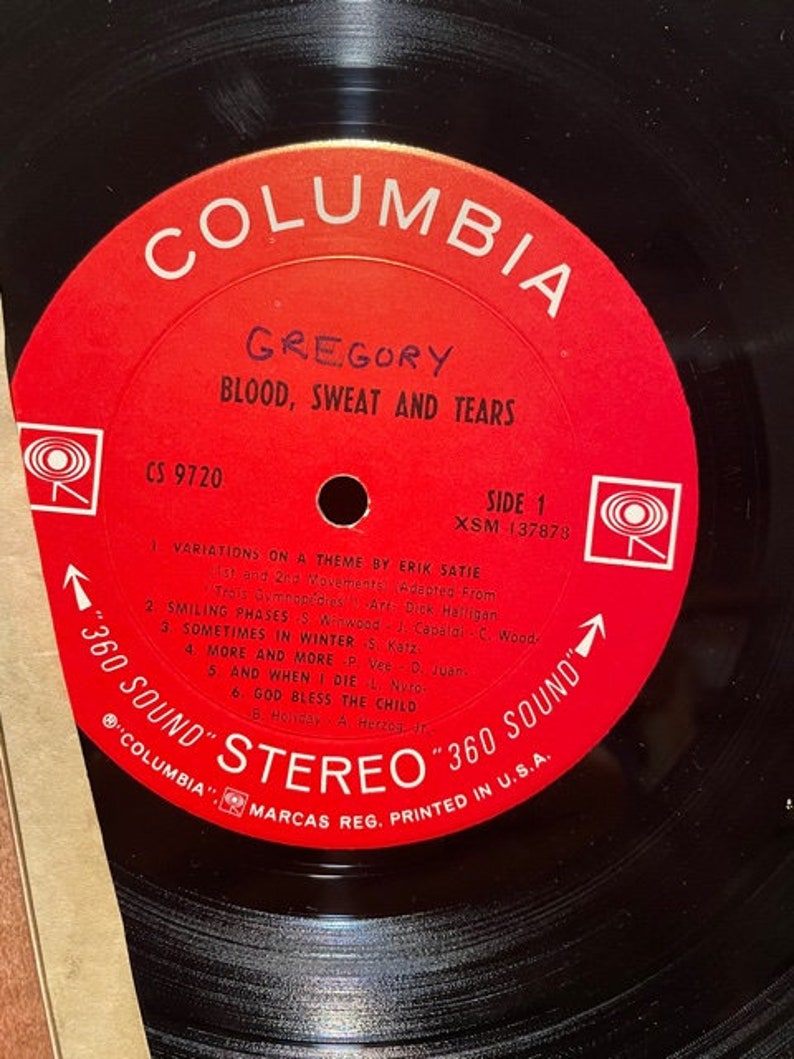 May include: A red vinyl record with the text "Columbia" in white, "Gregory" in black, and "Blood, Sweat and Tears" in white. The record is labeled "CS 9720" and "Side 1" with the song titles "Variations on a Theme by Erik Satie", "Smiling Phases", "Sometimes in Winter", "More and More", "And When I Die", and "God Bless the Child".