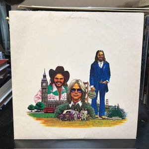 May include: A square vinyl record cover featuring a white background with a colorful illustration of three people, Big Ben, the Golden Gate Bridge, a red double-decker bus, and a purple vintage car on a grassy landscape.