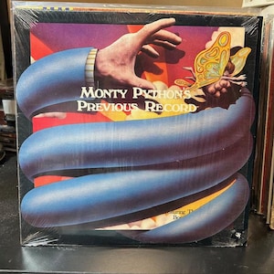 May include: A vinyl record album cover featuring the text "Monty Python's Previous Record". The artwork depicts a blue snake-like form with a hand reaching out, and a butterfly with a small figure. The background has purple and red hues.