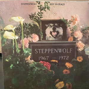 May include: Album cover for Steppenwolf's "Rest in Peace" featuring a memorial with a framed wolf portrait, surrounded by flowers. The band's name and years 1967-1972 are inscribed on a tombstone.