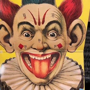 May include: A vintage carnival game target featuring a clown with a wide, toothy grin and a red tongue sticking out. The clown has red hair, blue hair, and a white ruffle collar. The target has numbers 10, 15, and 25 printed on it.